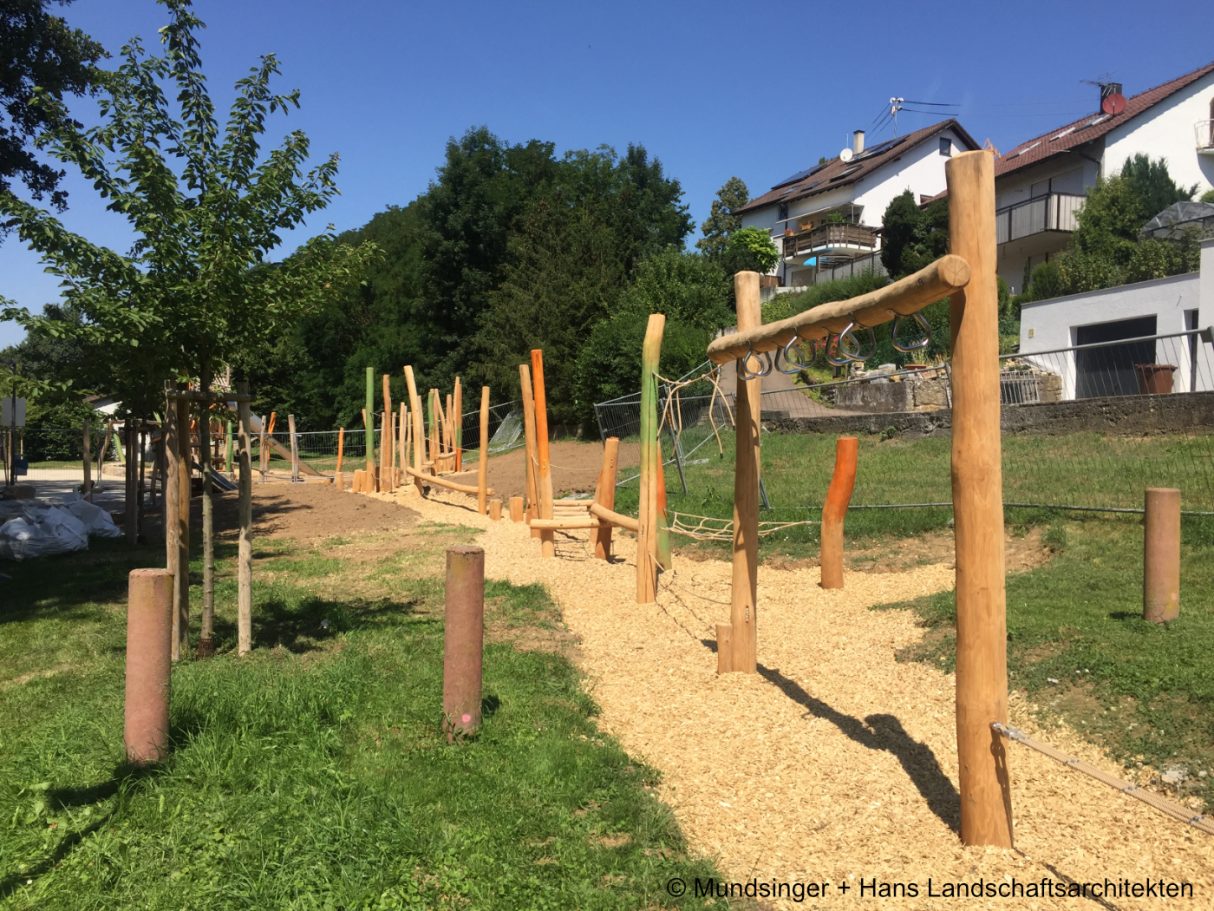 Ein Spielplatz mit Holzspielgeräten und einem Kiesweg, umgeben von Bäumen.