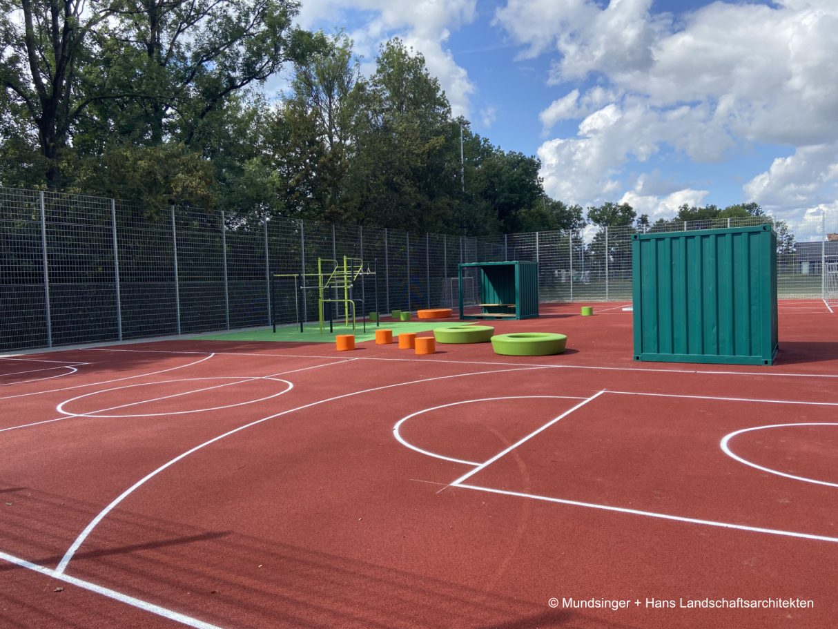 Spielplatz mit bunten Sitzgelegenheiten und grünen Containern auf einem roten Sportplatz.