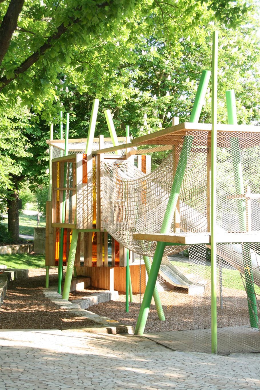 Spielplatz mit Klettergerüst aus Holz und Netz, umgeben von Bäumen.